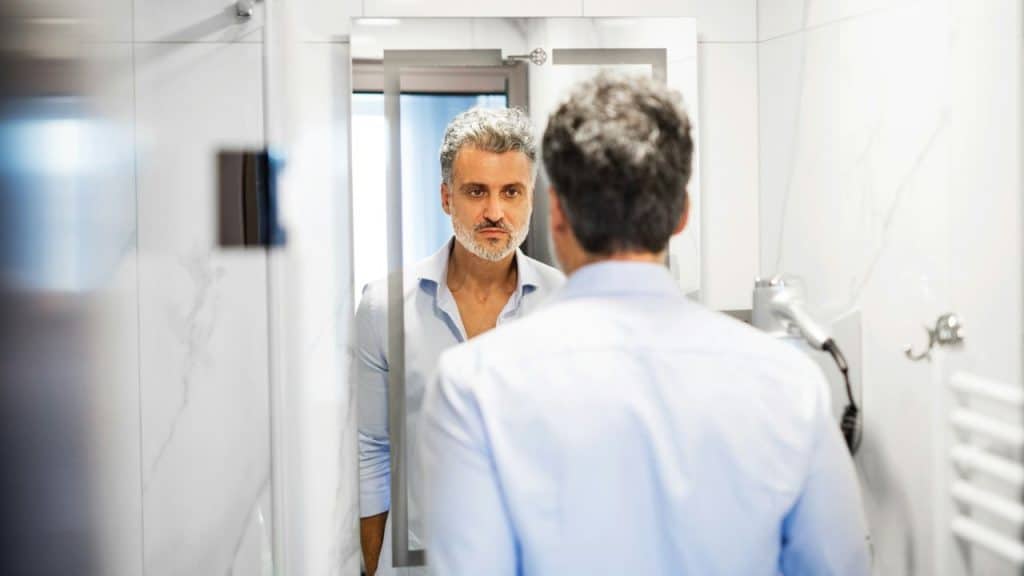 A man with gray hair looks at his reflection in a bathroom mirror with marble walls.