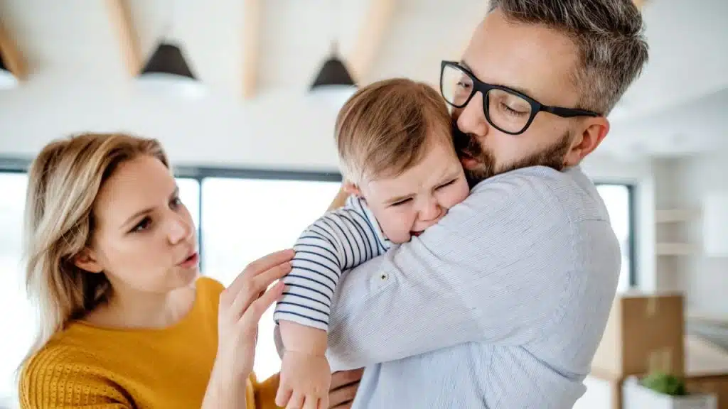 A man in a striped shirt holds a crying baby while a woman watches from behind.