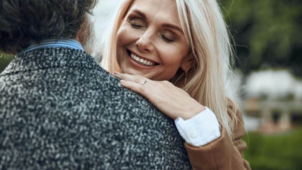 A smiling blonde woman rests her hand on the shoulder of a man from behind.