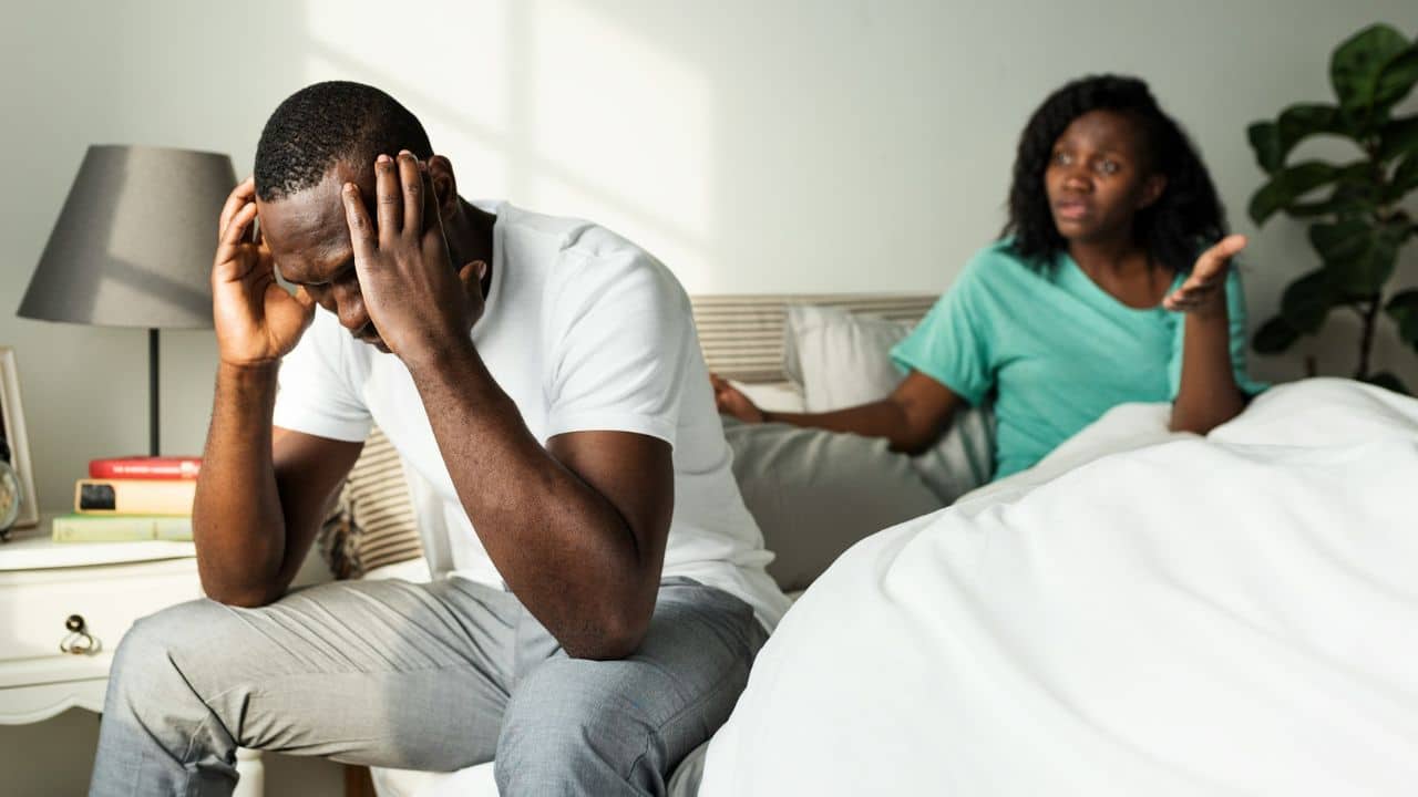 A man sits on a bed holding his head while a woman gestures behind him.