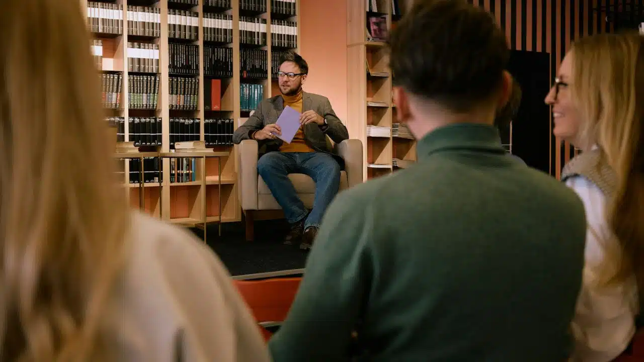 A man sits in an armchair holding a purple card while speaking to an audience.