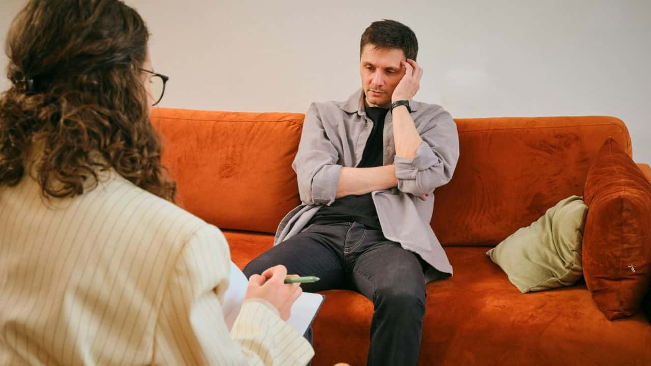 A man sits on an orange sofa while a woman takes notes during a session.