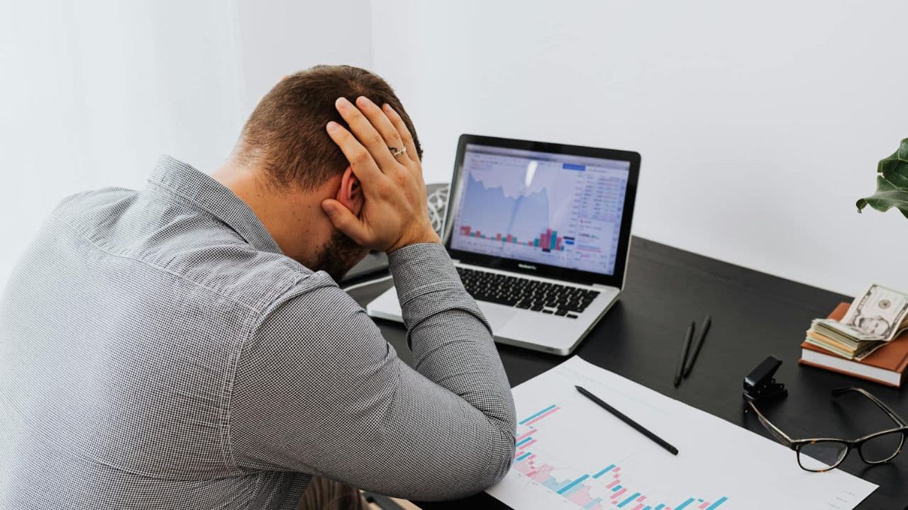 A man seen from behind sits at a desk with his hands on his head.
