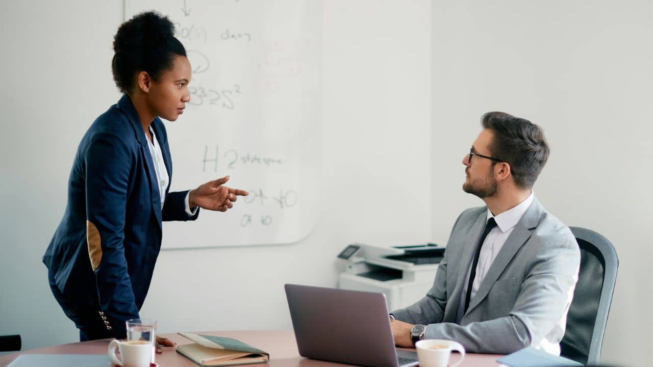 A woman in a suit stands and gestures toward a man seated at a desk.