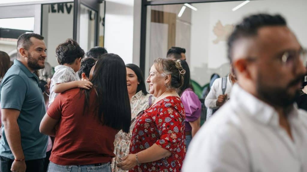 A group of people gather and talk indoors, including a smiling woman in a floral shirt.