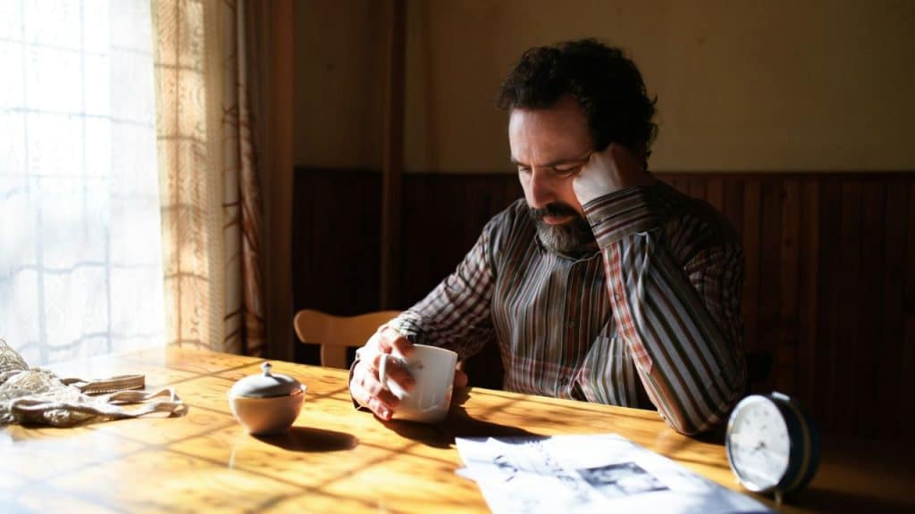 A bearded man in a striped shirt sits at a table, resting his head on his hand.