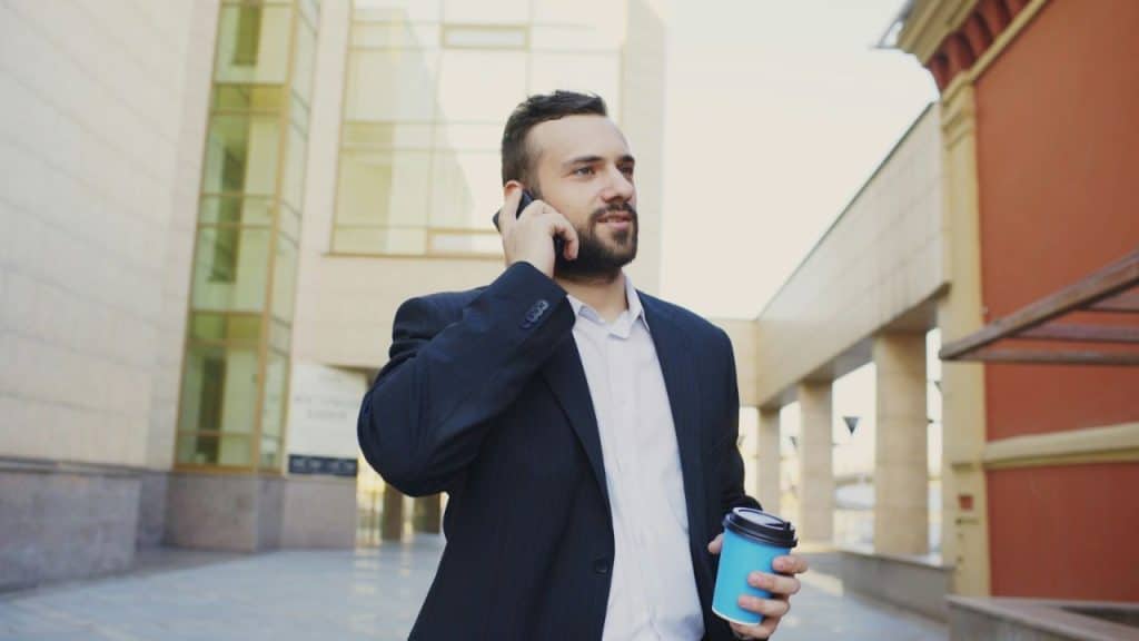 A man in a suit jacket talks on a phone while holding a blue cup.