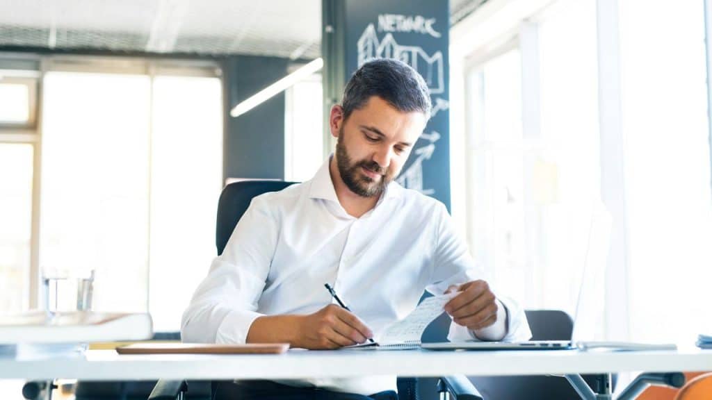 A bearded man in a white dress shirt sits at a desk, writing in a notebook.