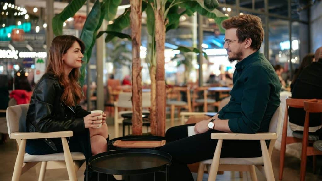 A man and woman sit in chairs at a cafe table while holding white mugs.