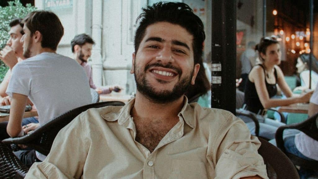 A smiling man with a dark beard sits outdoors at a table with people behind him