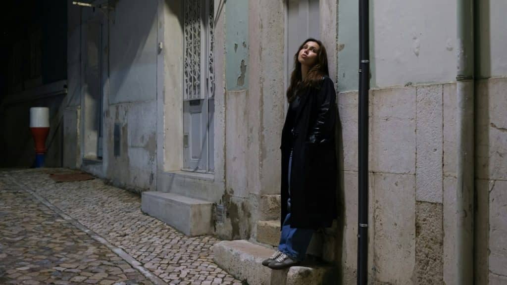 A woman in a black coat leans against a wall in a narrow alley at night.