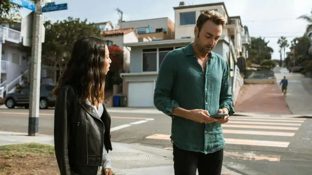 A man looks down at his phone while a woman stands beside him on a residential street.