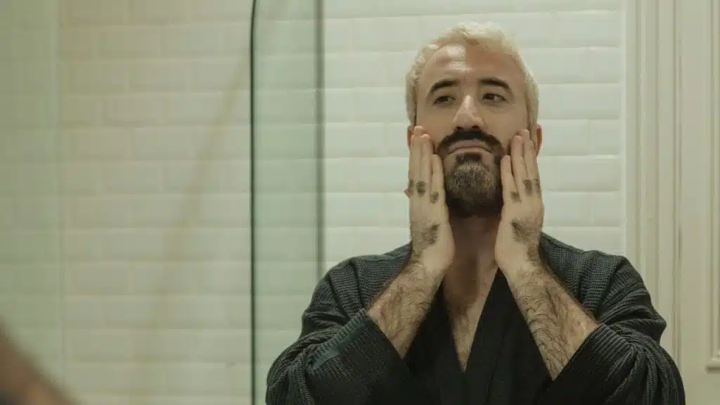 A man with blonde hair and a dark beard touches his face in a mirror.