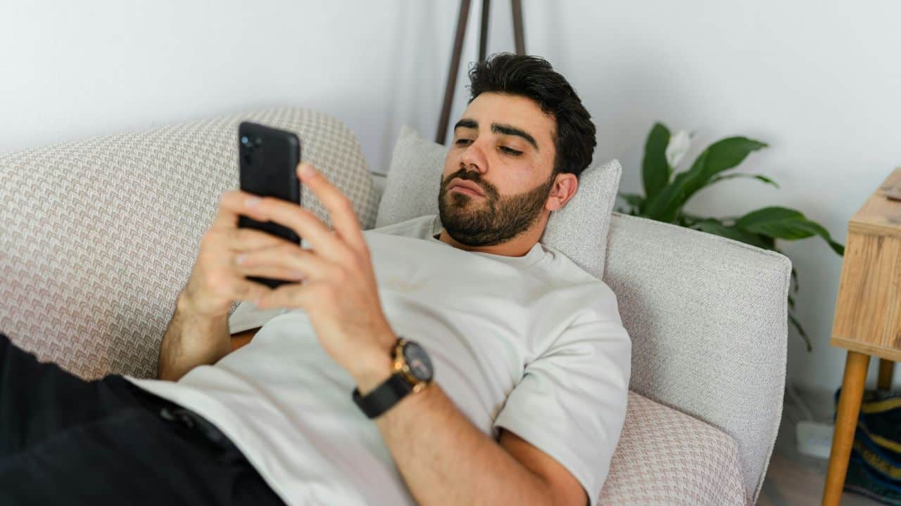 A bearded man lies on a light-colored sofa while looking down at his smartphone.