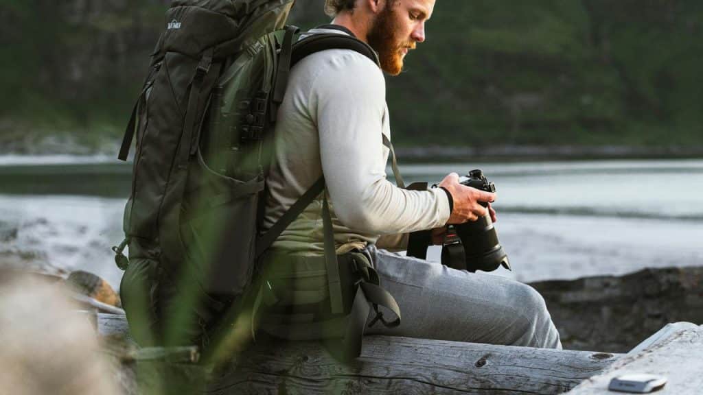 A man with a backpack sitting outdoors holding a camera.