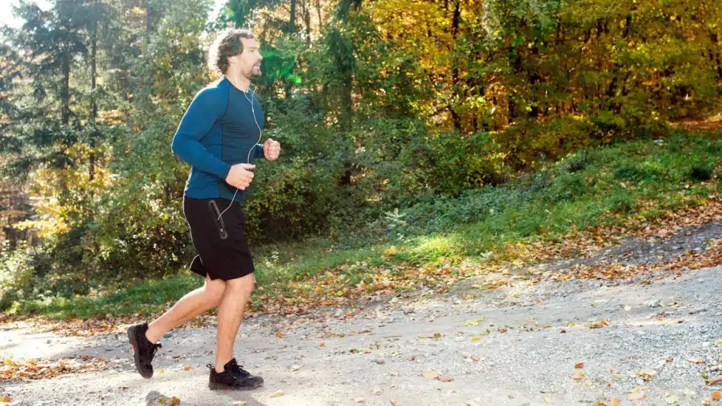 A man in athletic gear jogs along a leaf-covered path through a sunlit forest.