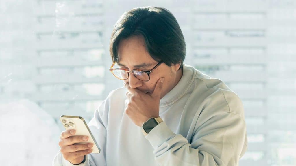 A man wearing glasses and a light-colored sweatshirt looks at his smartphone while resting his chin on his hand.