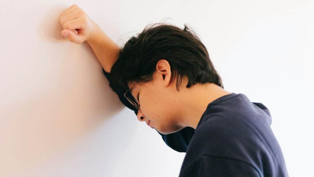 A person with glasses leans their forehead and arm against a white wall.