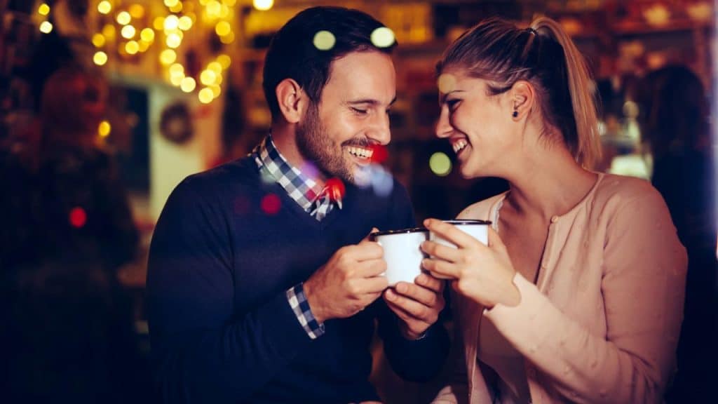A man and woman laugh together while holding mugs in a dimly lit, festive setting.