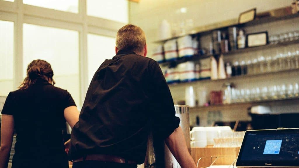 Two people in dark shirts stand with their backs to the camera in a cafe.