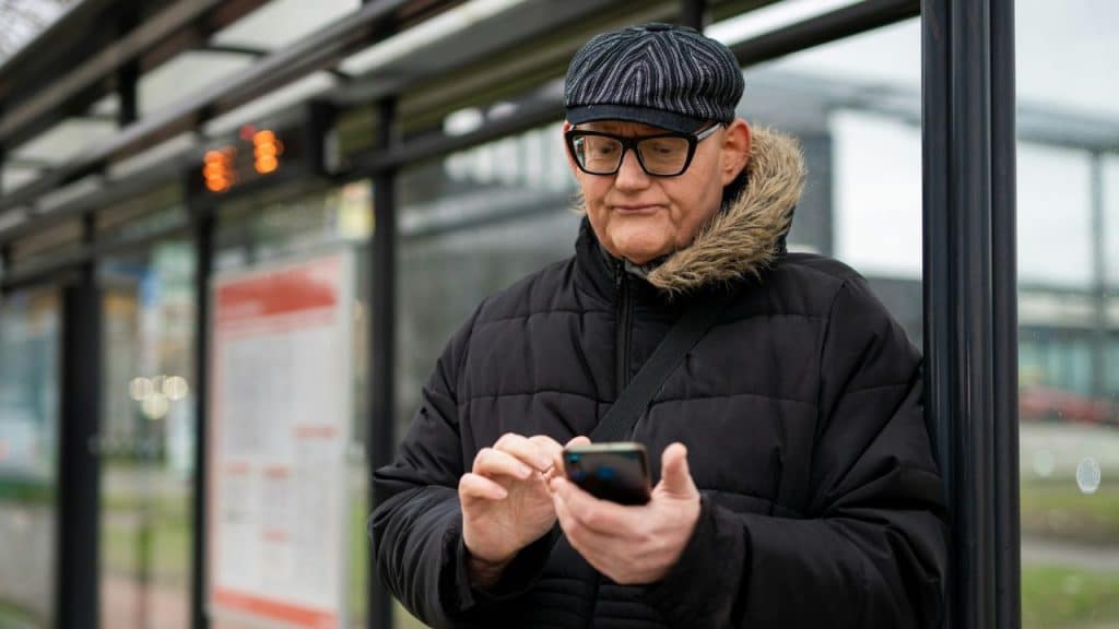 A man in a flat cap and glasses looks down at his phone while outdoors.