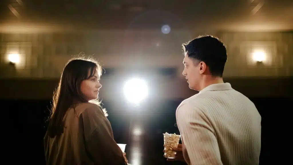 A man and a woman face each other in a dimly lit theater holding popcorn.
