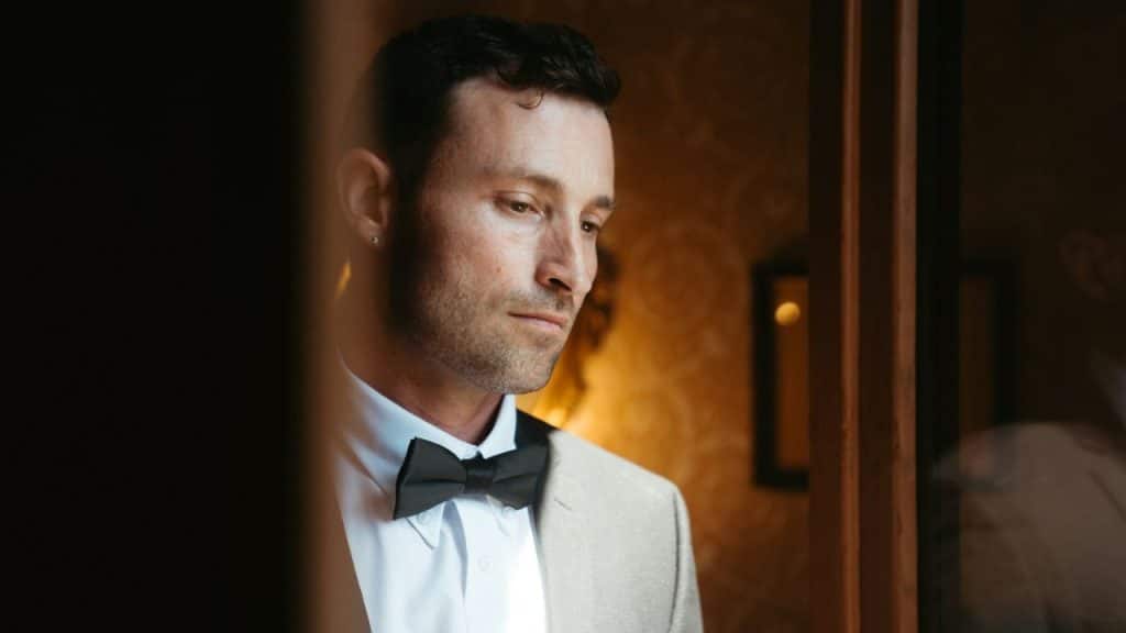 A man in a tan tuxedo jacket and black bowtie looks downward in a dimly lit room.