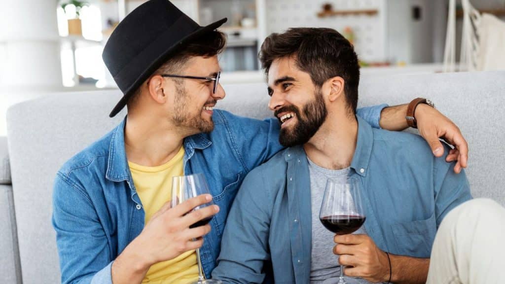 Two men in casual shirts sit together on a couch, smiling and holding wine glasses.