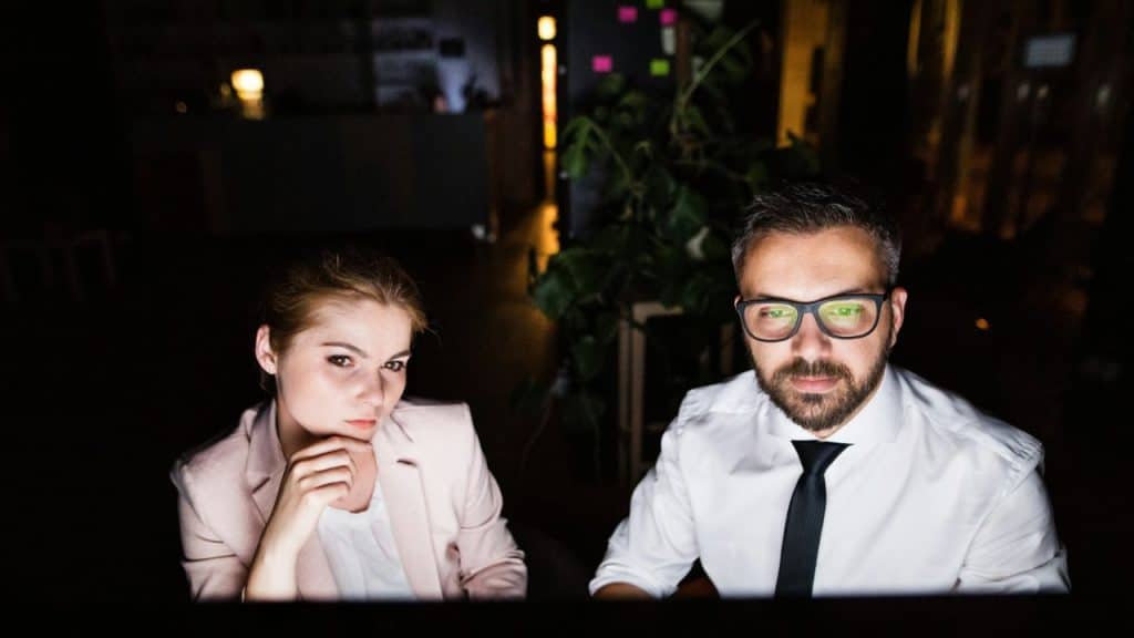 A man and woman in business attire look toward a glowing screen in the dark.