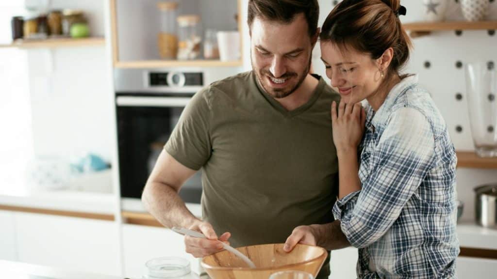 A man and woman stand in a kitchen while the man stirs a wooden bowl.