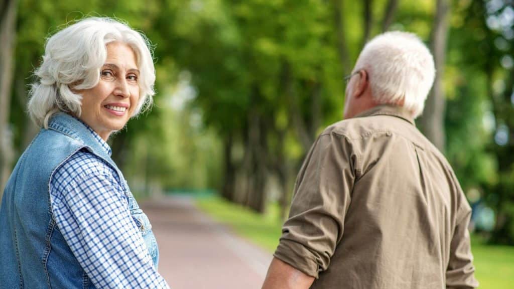 An older woman with white hair smiles at the camera while walking on a path.