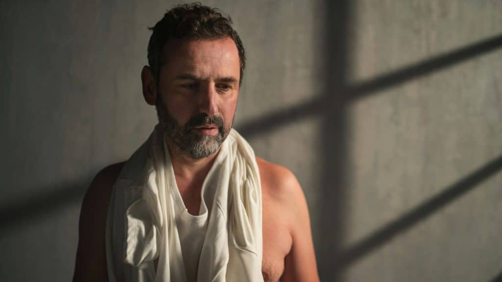 A shirtless man with a white shirt draped around his neck stands against a wall.