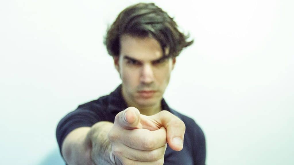 A man in a black polo shirt points his finger directly toward the camera lens.
