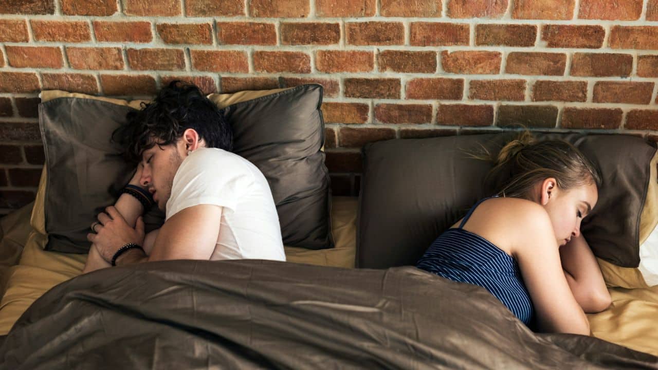 A man and woman lie in bed facing away from each other with space between them.