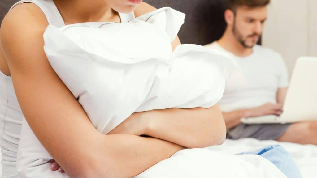 A woman hugging a pillow while a man works on a laptop in the background.