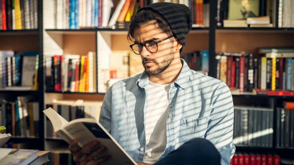 A man in glasses and a beanie reading a book in a library.