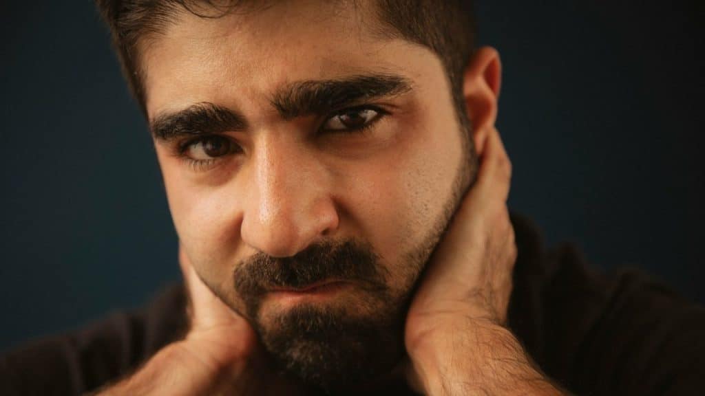 A bearded man with an intense expression has his hands clasped behind his neck.