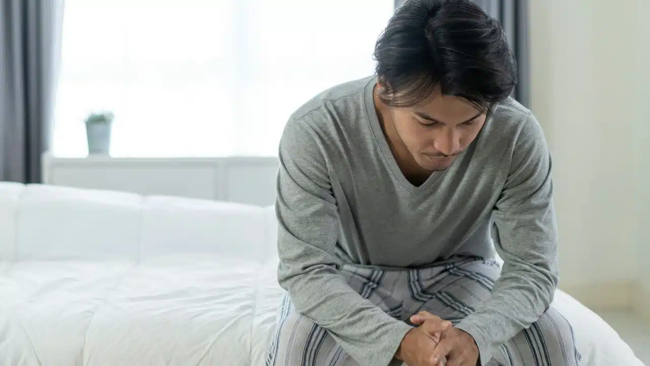A man in pajamas sits on the edge of a bed with his head bowed down.