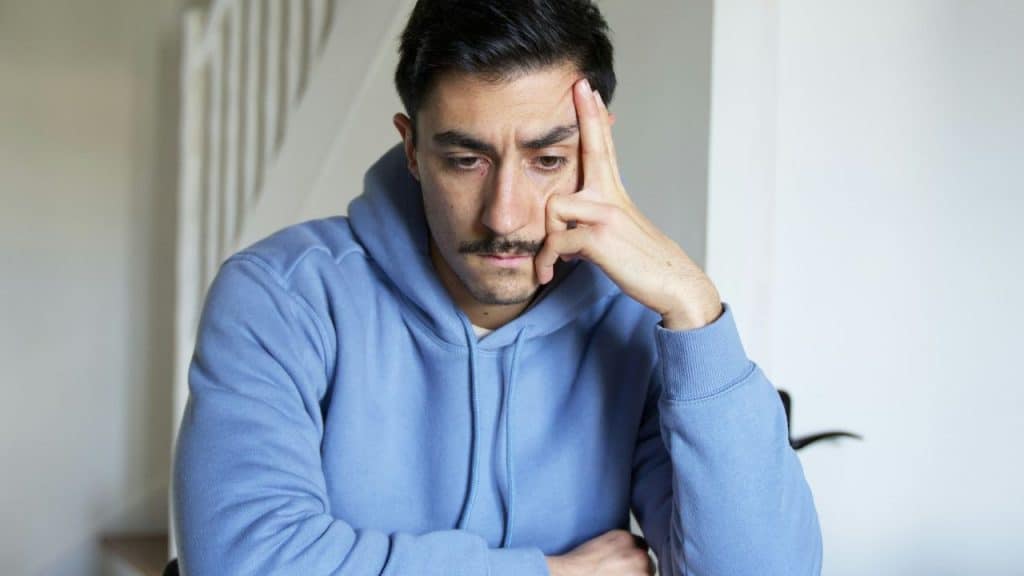 A man in a blue hoodie rests his head on his hand with a tired expression.
