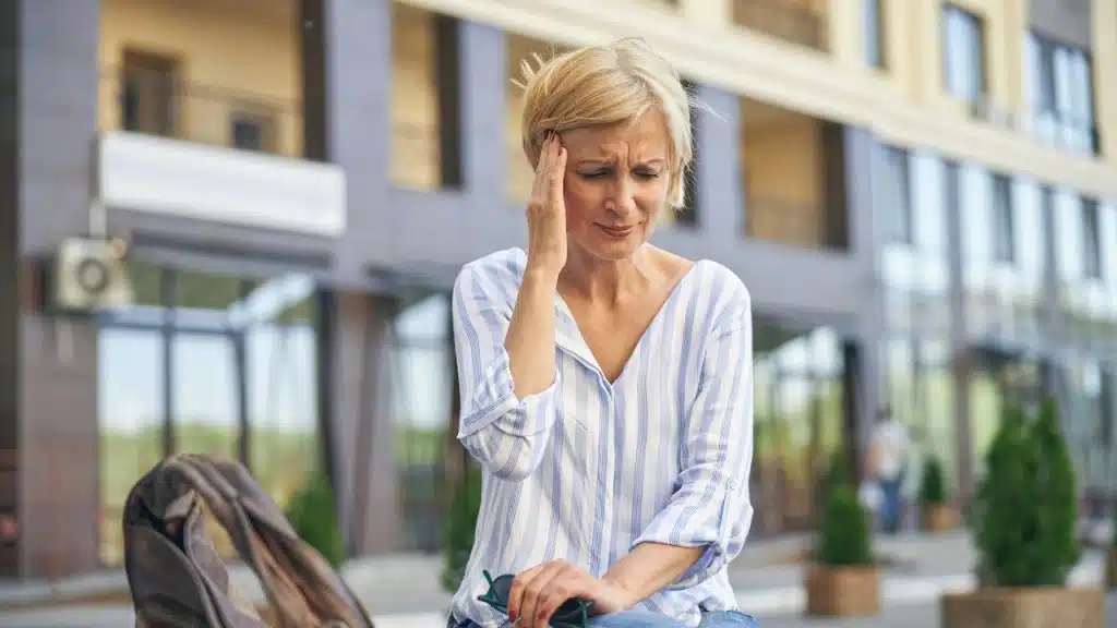 A woman with short blonde hair sits on an outdoor bench, pressing her hand to her temple with a distressed expression.