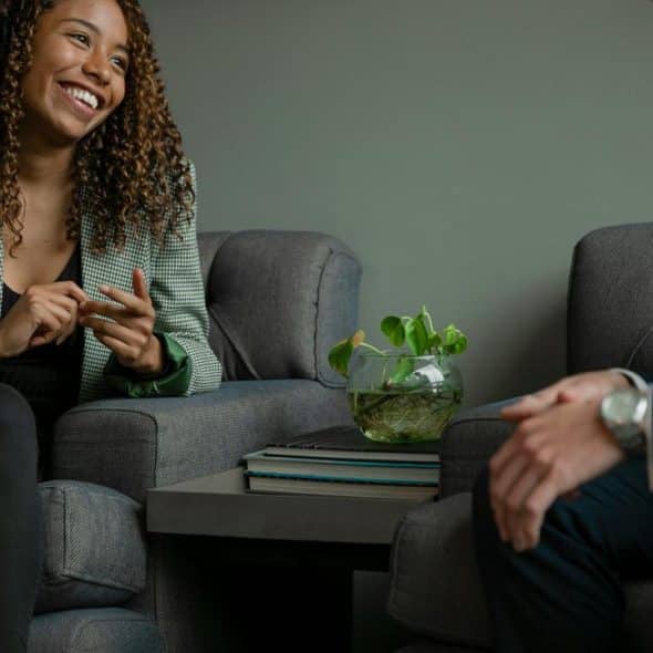 A smiling woman chatting with a colleague while seated in a modern office lounge.