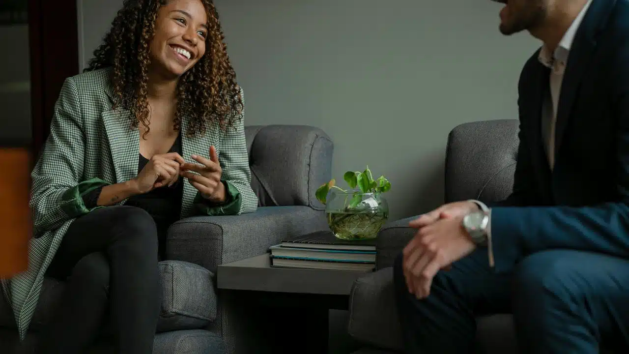 A smiling woman chatting with a colleague while seated in a modern office lounge.