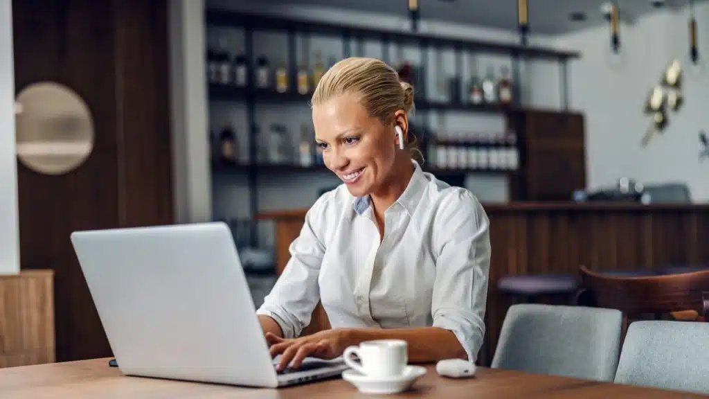 A blonde woman wearing wireless earbuds smiles while typing on a laptop in a cafe.