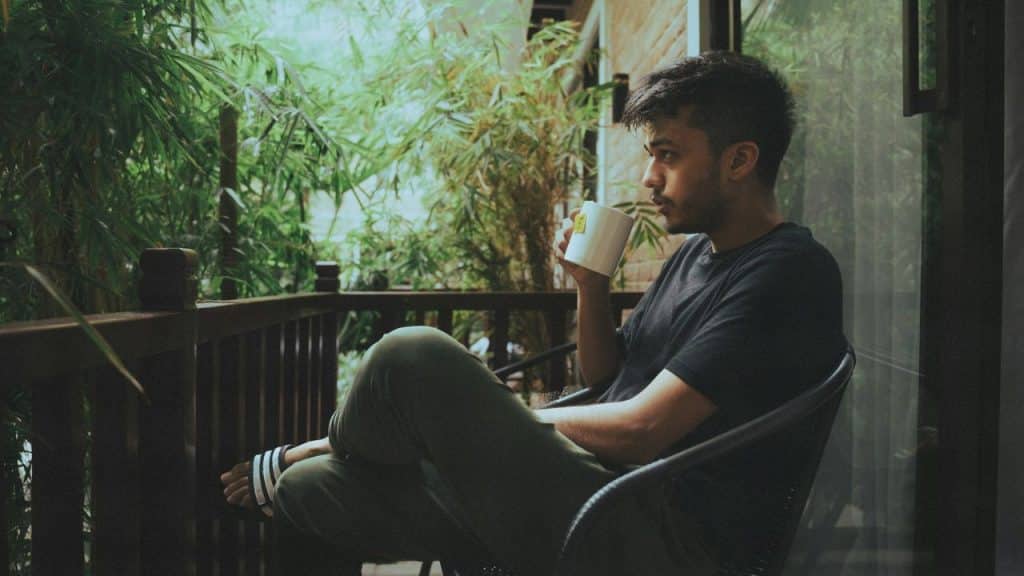 A man sitting on a balcony holding a mug and looking at green foliage.