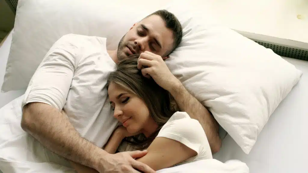 A man and woman lie in bed together on white pillows with their eyes closed peacefully.