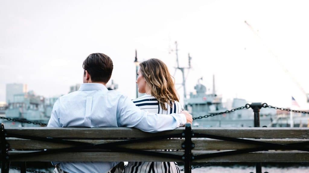 A couple sitting on a bench by the water, looking out at ships together.