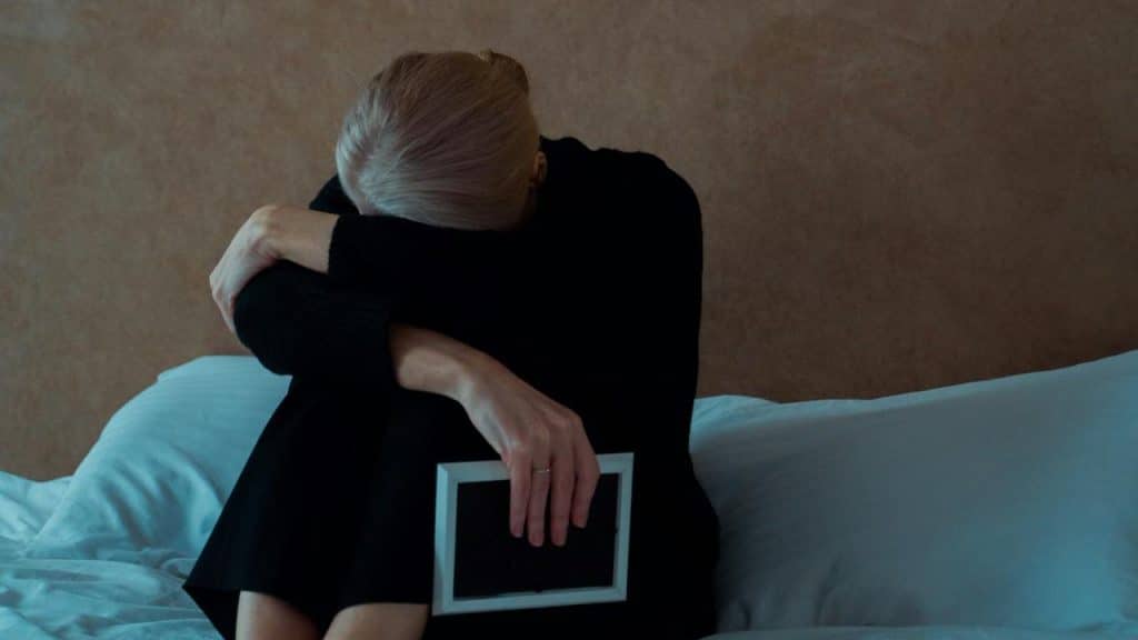 A person dressed in black sits on a bed with their head resting on their arms while holding an empty white picture frame.