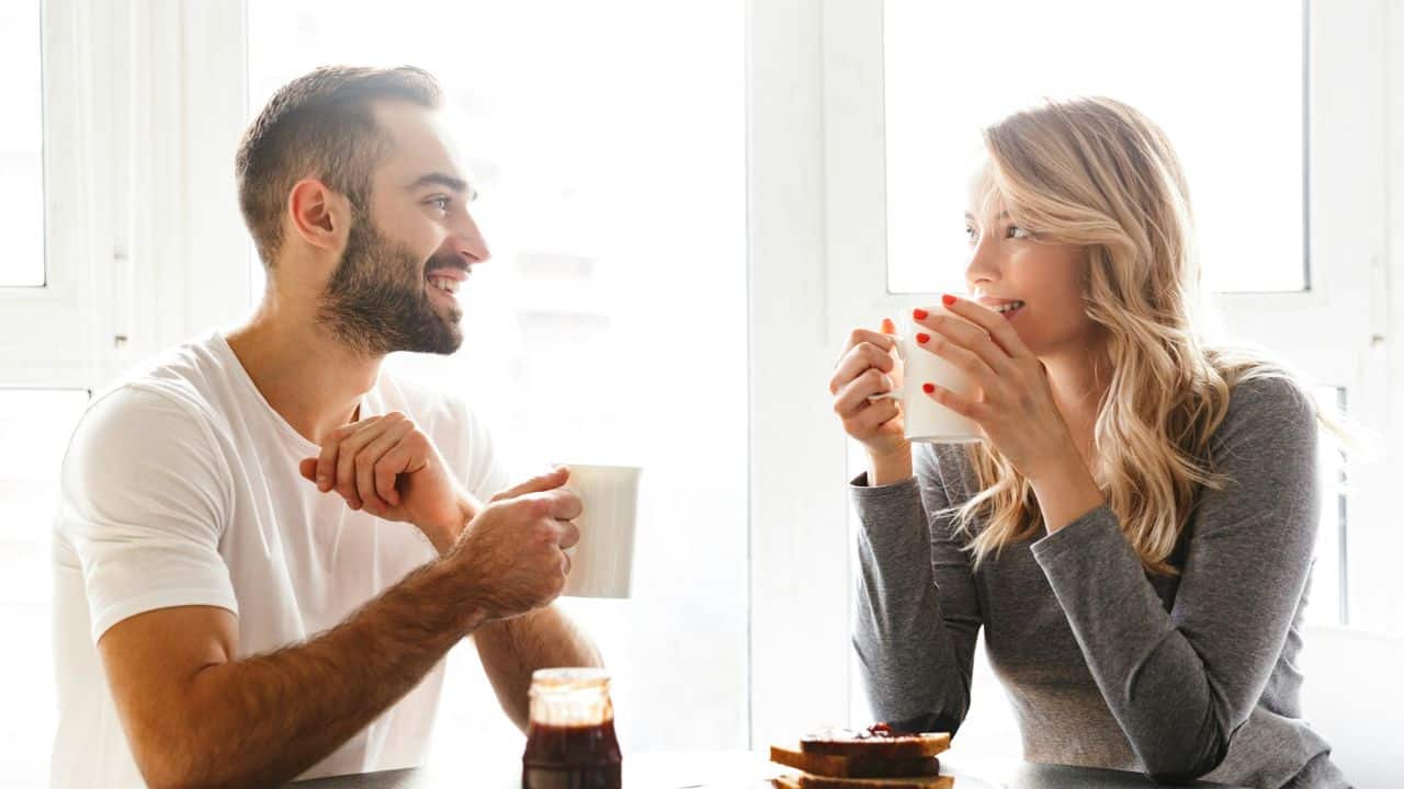 A smiling man and woman sit at a table holding mugs while looking at each other.