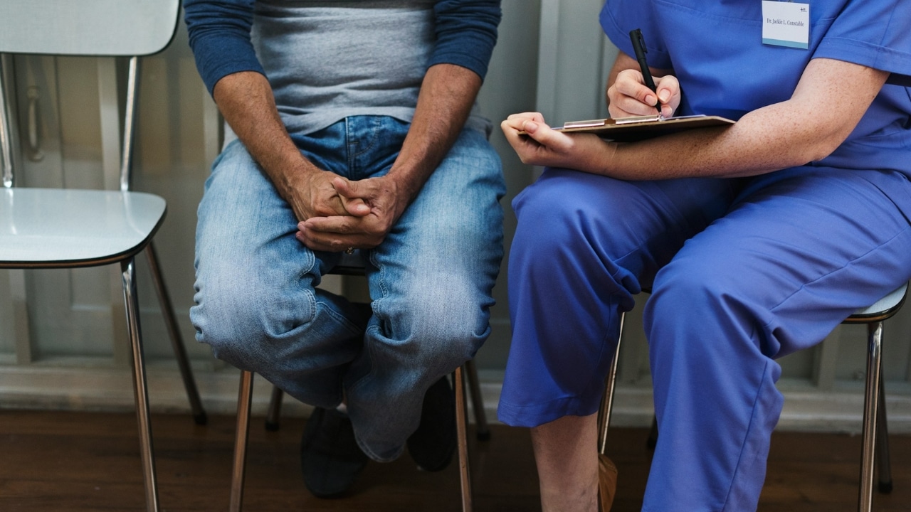 A healthcare worker in blue scrubs writes on a clipboard next to a seated patient.