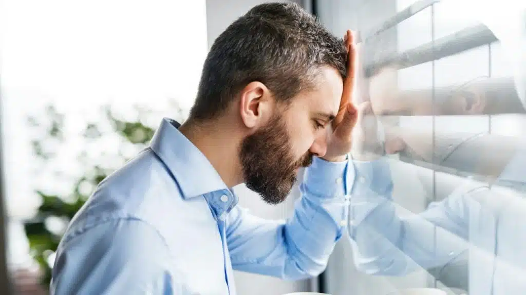 A stressed man in a blue shirt leaning his forehead against a window with his eyes closed.